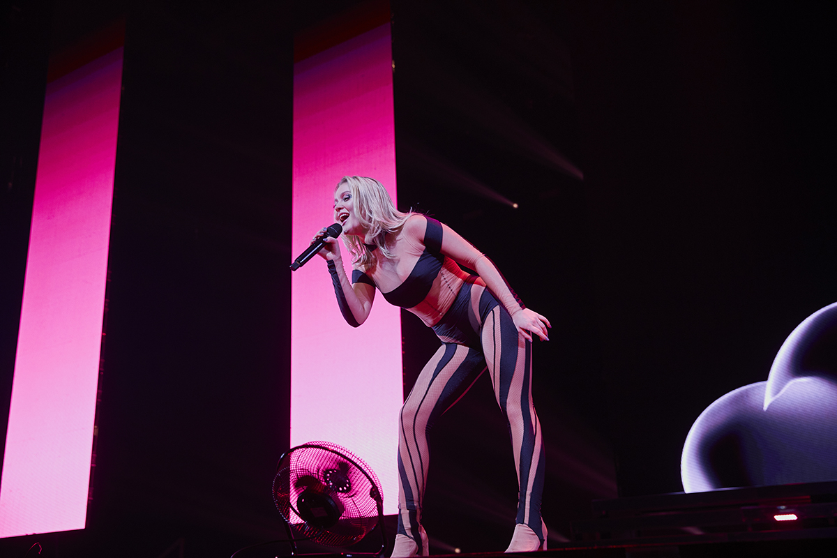 Zara Larsson står i mitten av bilden och sjunger. Hon böjer sig lite ner framåt. Det är rosa bakgrundsljus bakom henne. 