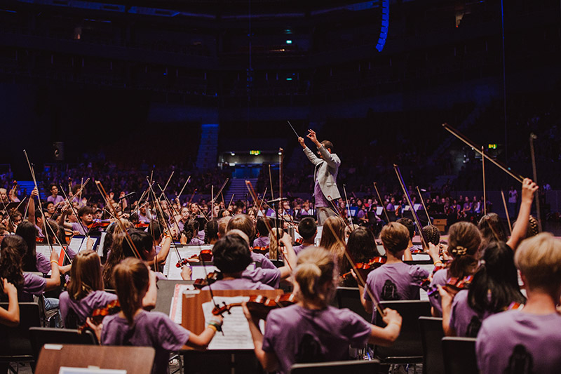 En dirigent leder en stor orkester med barn och unga.