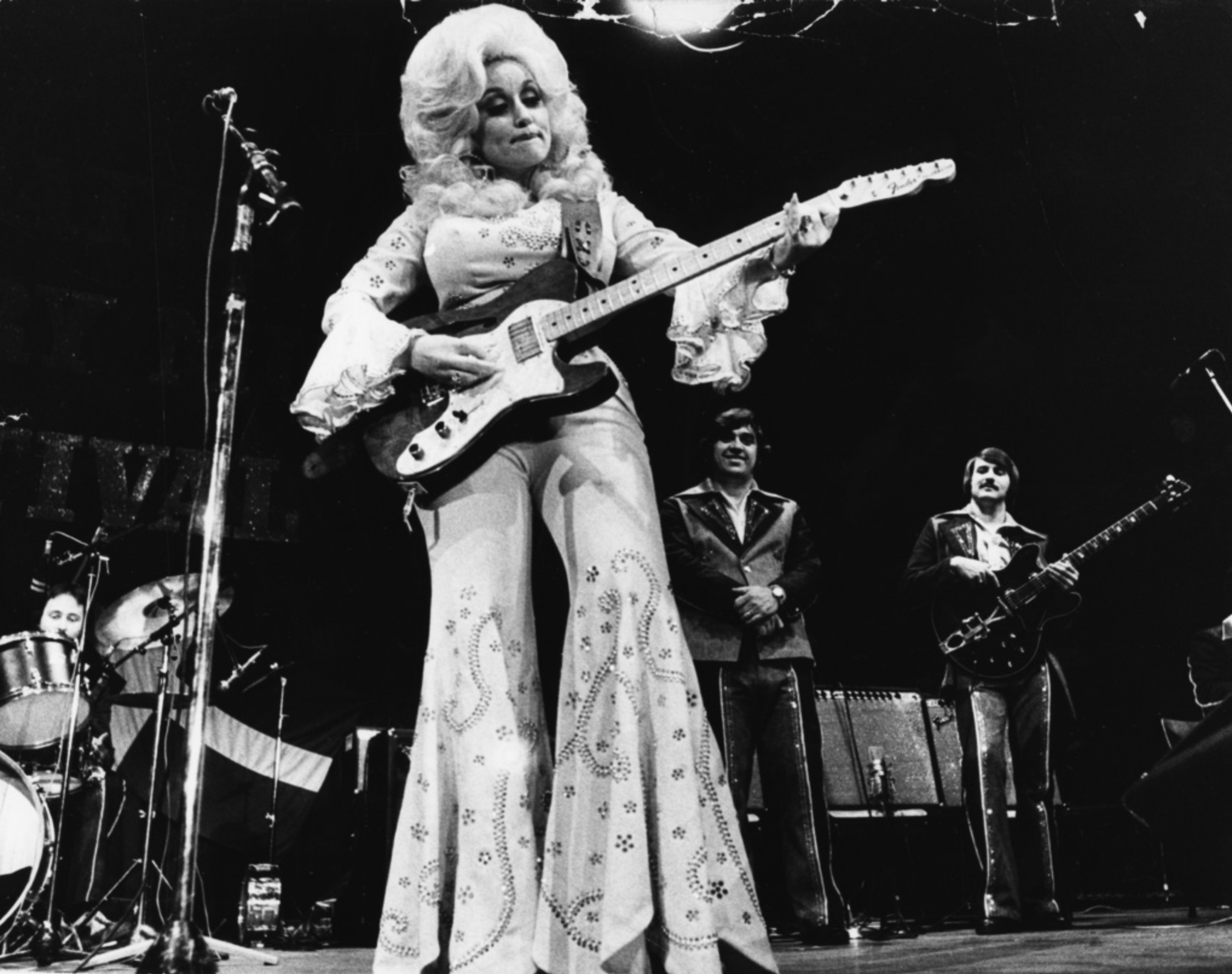 Dolly Parton med en gitarr och i en byxdress med utsvängda ben.