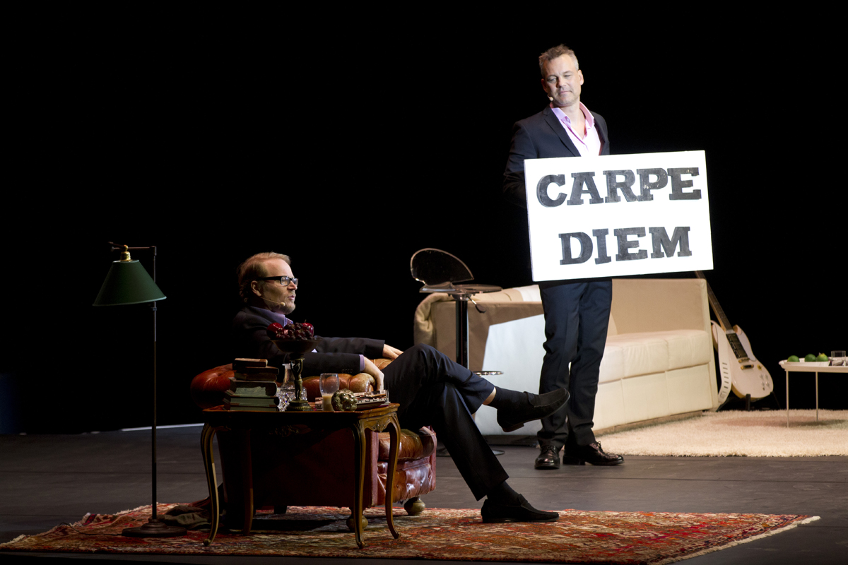 Fredrik Lindström sitter i en fåtölj och Henrik Schyffert håller en skylt som det står Carpe Diem på.
